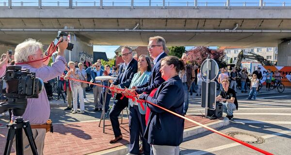Mehrere Personen halten ein rotes Band und schneiden es durch, im Hintergrund eine Brücke und Menschen